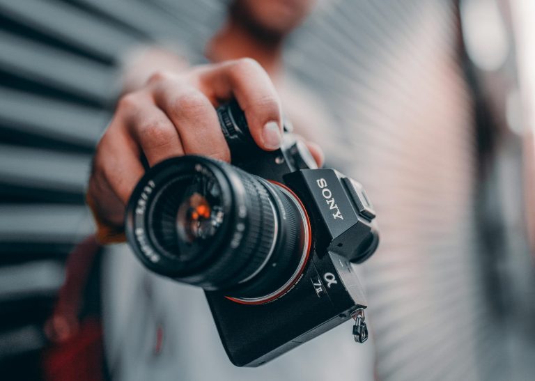 A person extends a Canon camera towards the viewer in an outdoor setting.